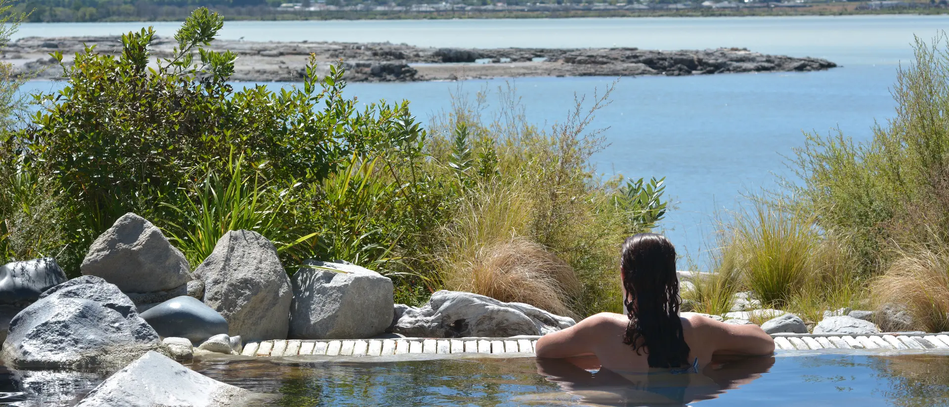 A person relaxes in a hot spring, surrounded by rocks and greenery, with a serene lake and mountains in the background.