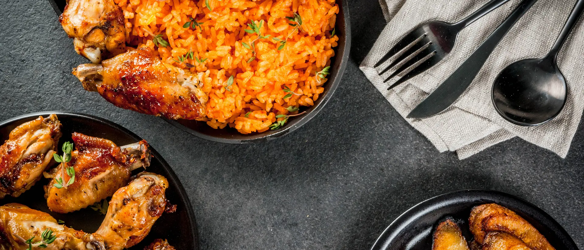 Two bowls of chicken wings and rice, accompanied by fried plantains and utensils on a dark tabletop.