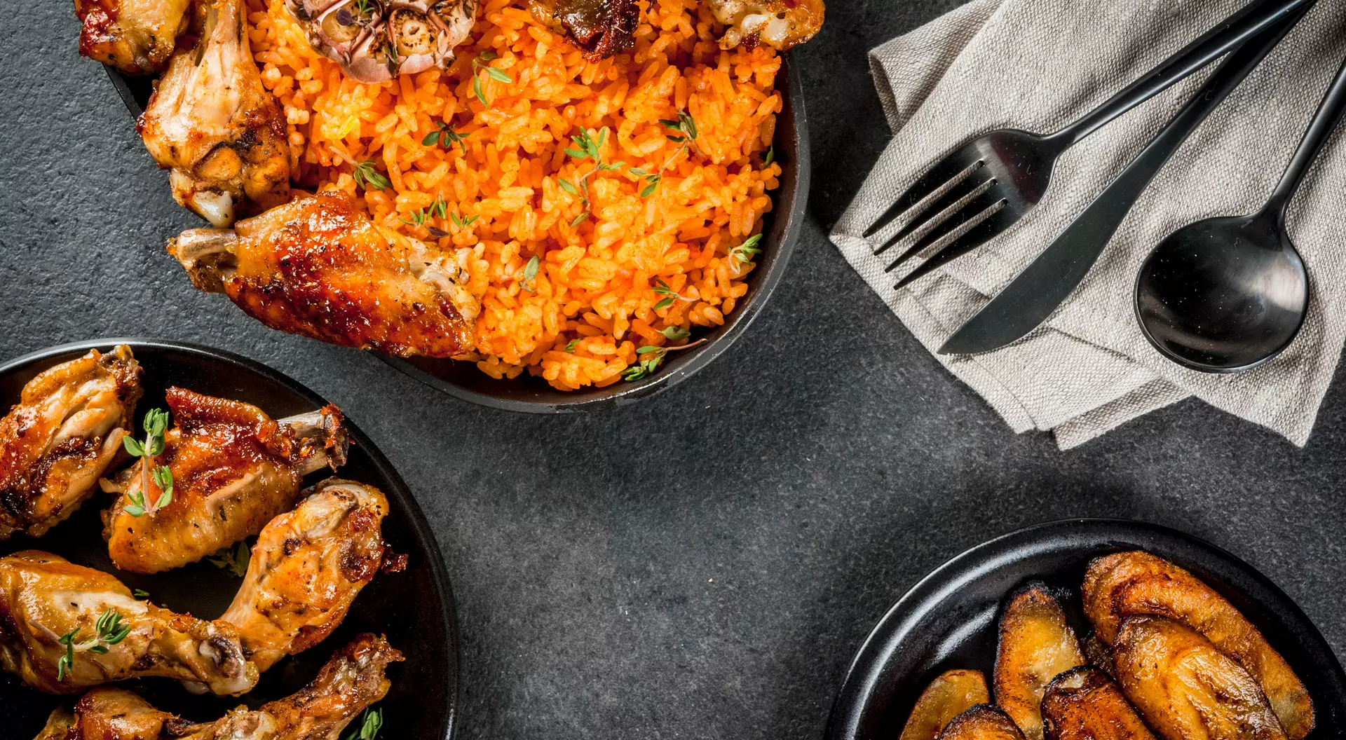 Two bowls of chicken wings and rice, accompanied by fried plantains and utensils on a dark tabletop.