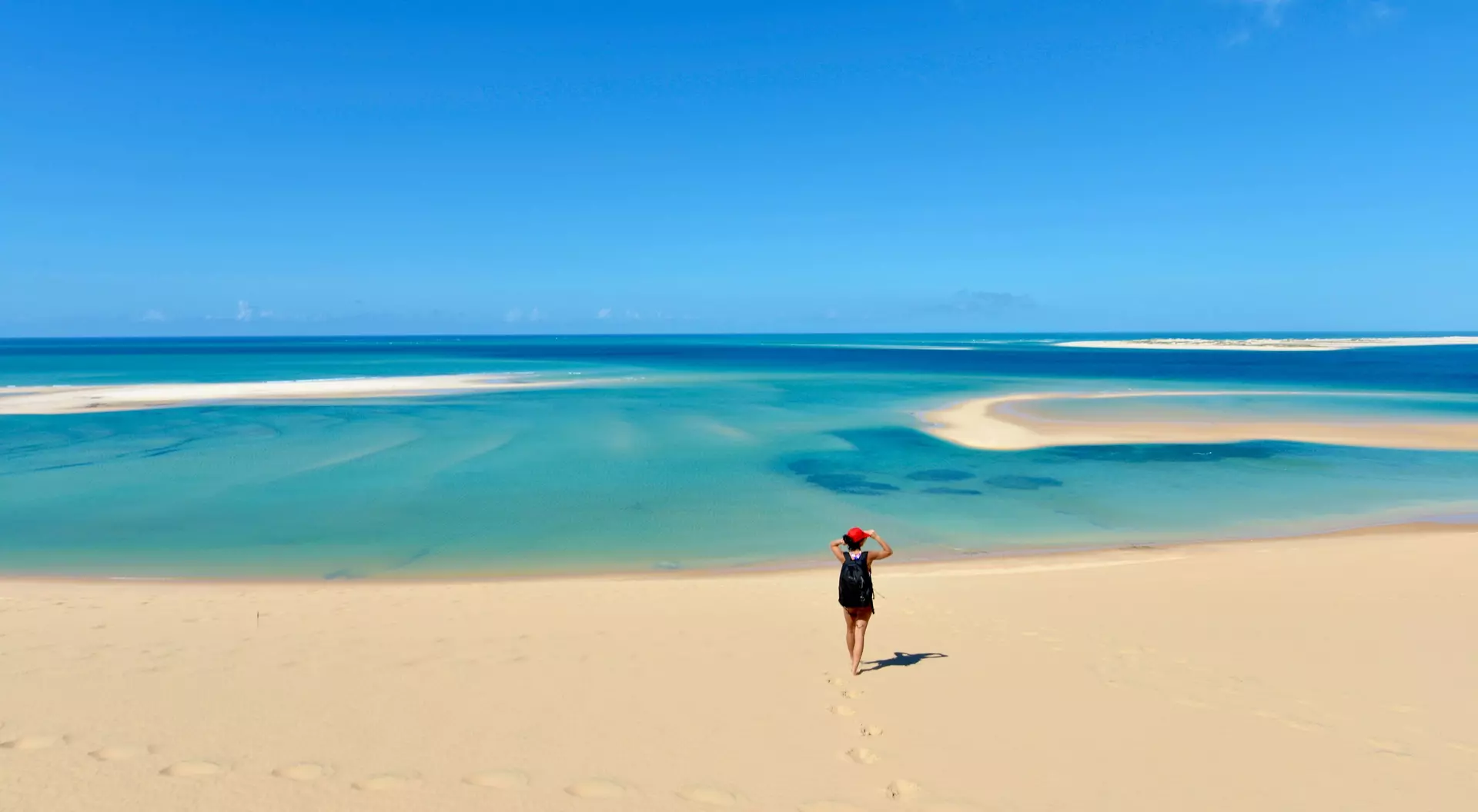 A person walks along a sandy beach, with clear blue water and a bright sky in the background.