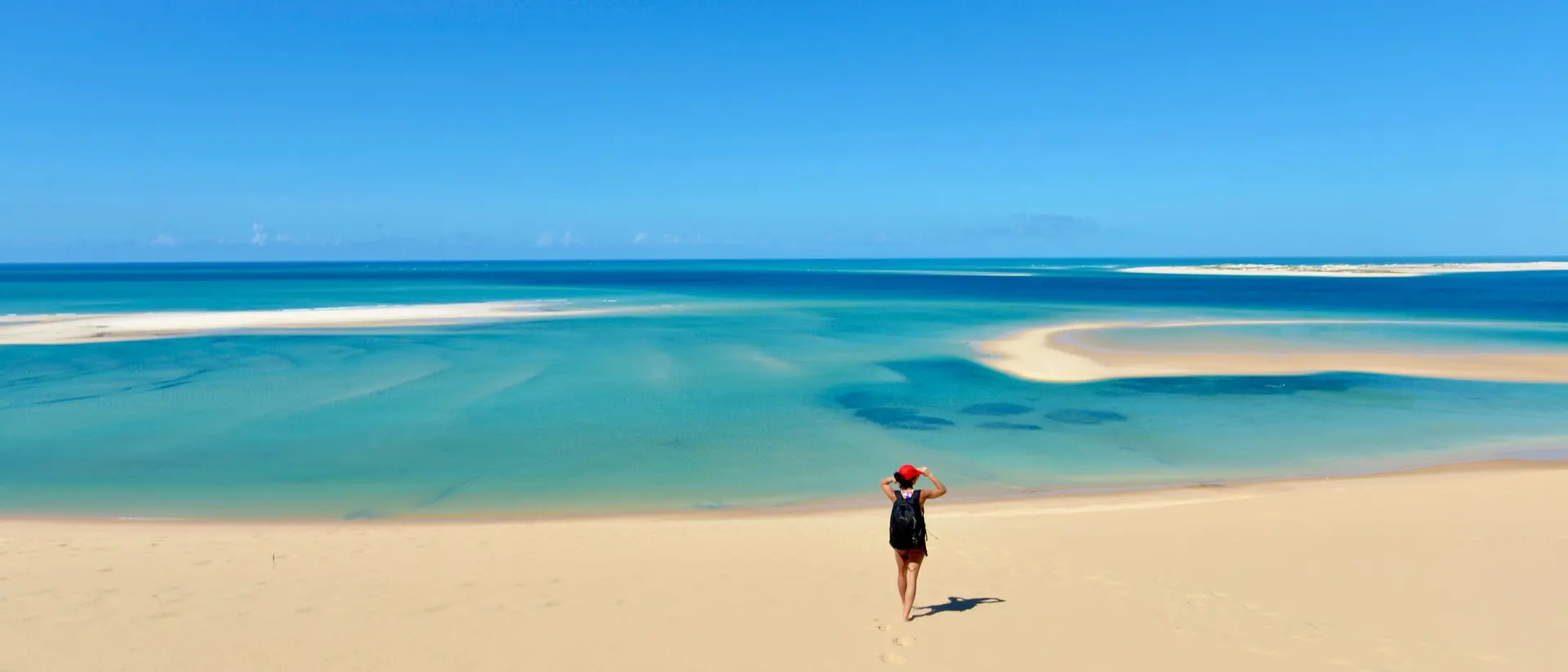 A person walks along a sandy beach, with clear blue water and a bright sky in the background.