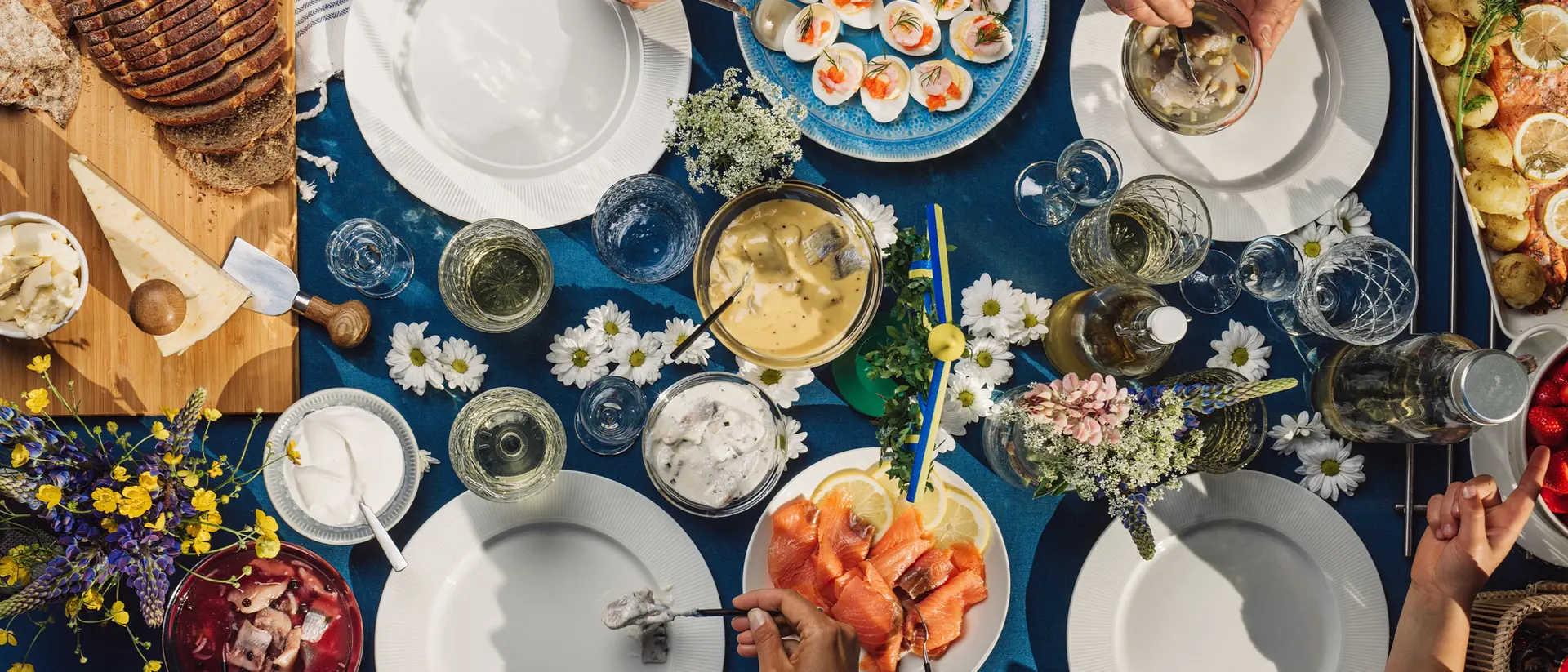 Aerial view of a vibrant outdoor table set with various dishes, drinks, and floral decorations, with hands reaching for food.