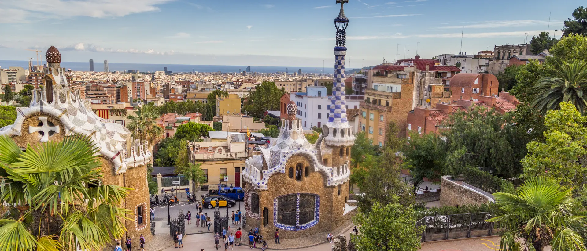 A panoramic view of Park Güell in Barcelona, featuring colorful buildings, palm trees, and a distant view of the city.