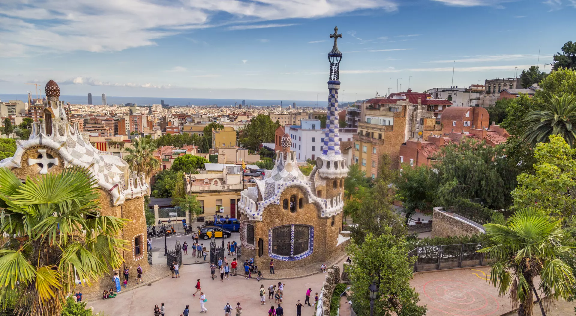 A panoramic view of Park Güell in Barcelona, featuring colorful buildings, palm trees, and a distant view of the city.