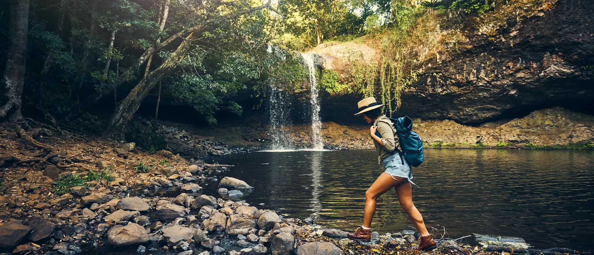 A person in shorts and a hat walks along rocks by a forest stream, with a waterfall in the background.