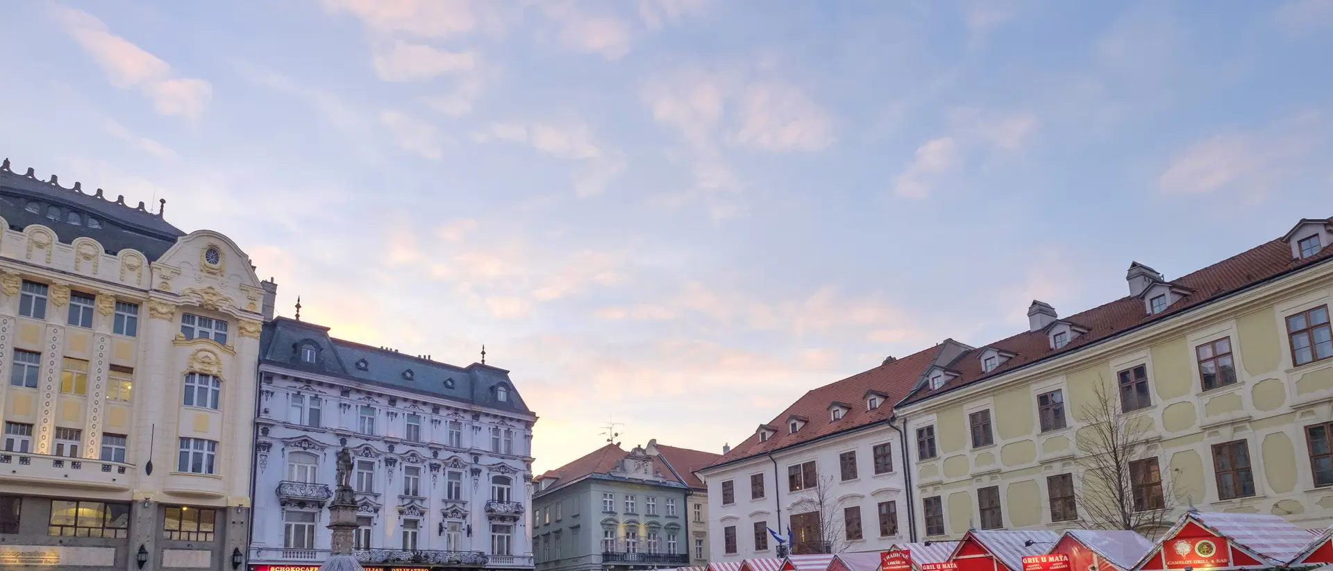 Crowded market scene with stalls under a colorful sky, surrounded by historic buildings.
