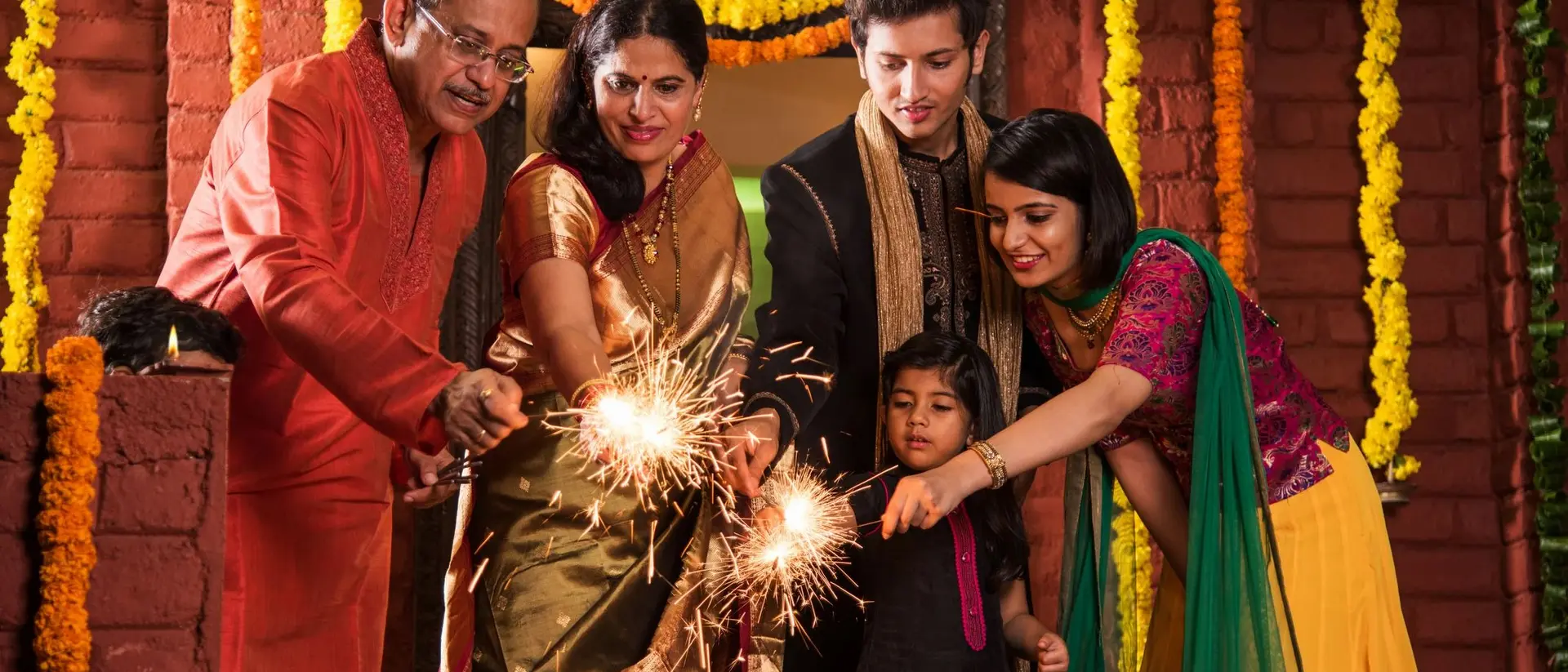 A family celebrates together, lighting sparklers in traditional attire surrounded by colorful flowers and festive decorations.