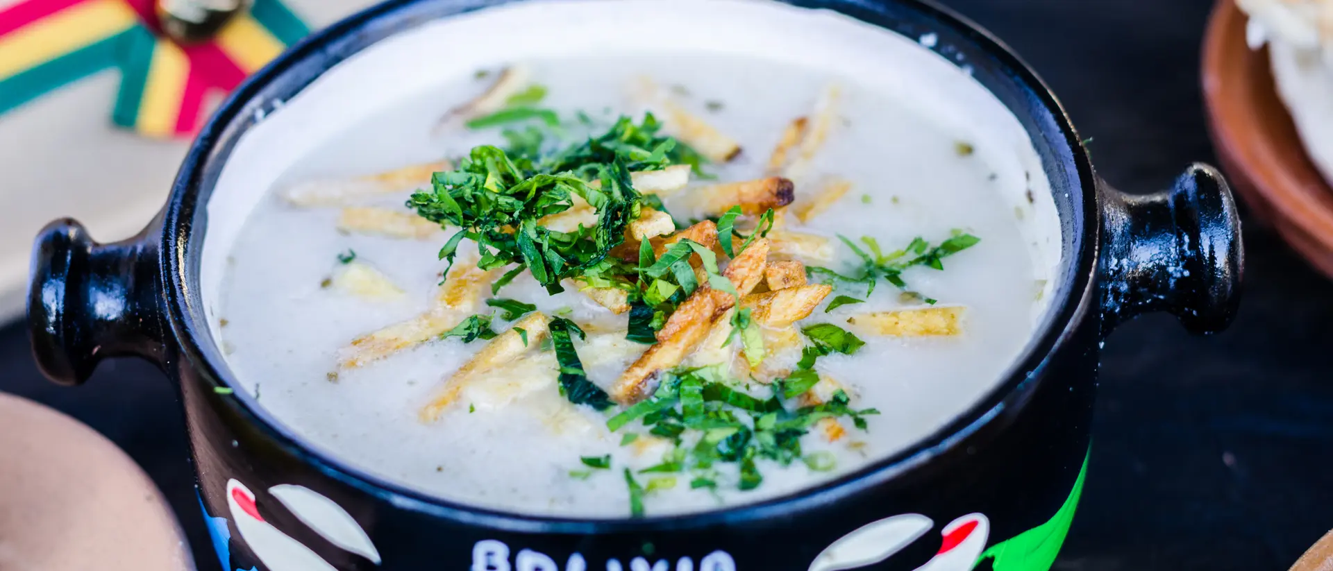 A colorful bowl of traditional Bolivian soup topped with herbs and crispy strips, alongside bread on a wooden surface.