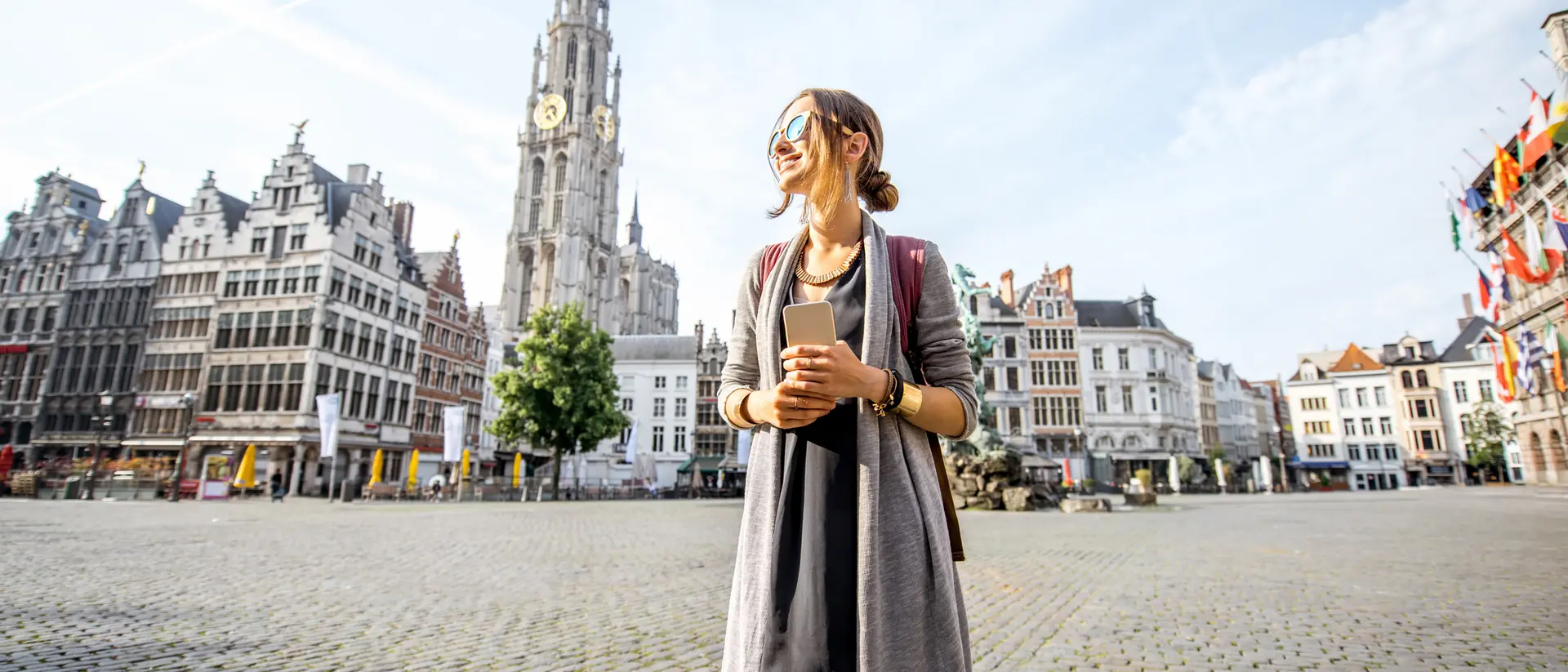 A woman stands in a plaza with historic buildings and a clock tower in the background, enjoying a sunny day.