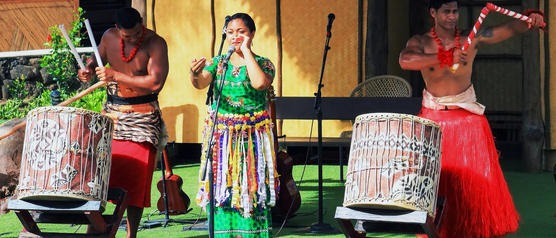 Three performers on stage: one singing, two drummers, dressed in traditional attire, with musical instruments and a vibrant backdrop.