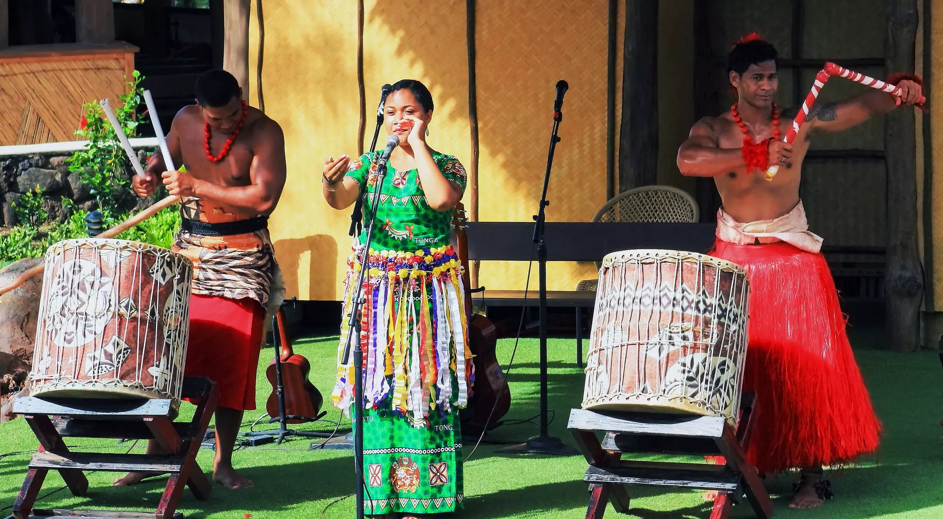 Three performers on stage: one singing, two drummers, dressed in traditional attire, with musical instruments and a vibrant backdrop.