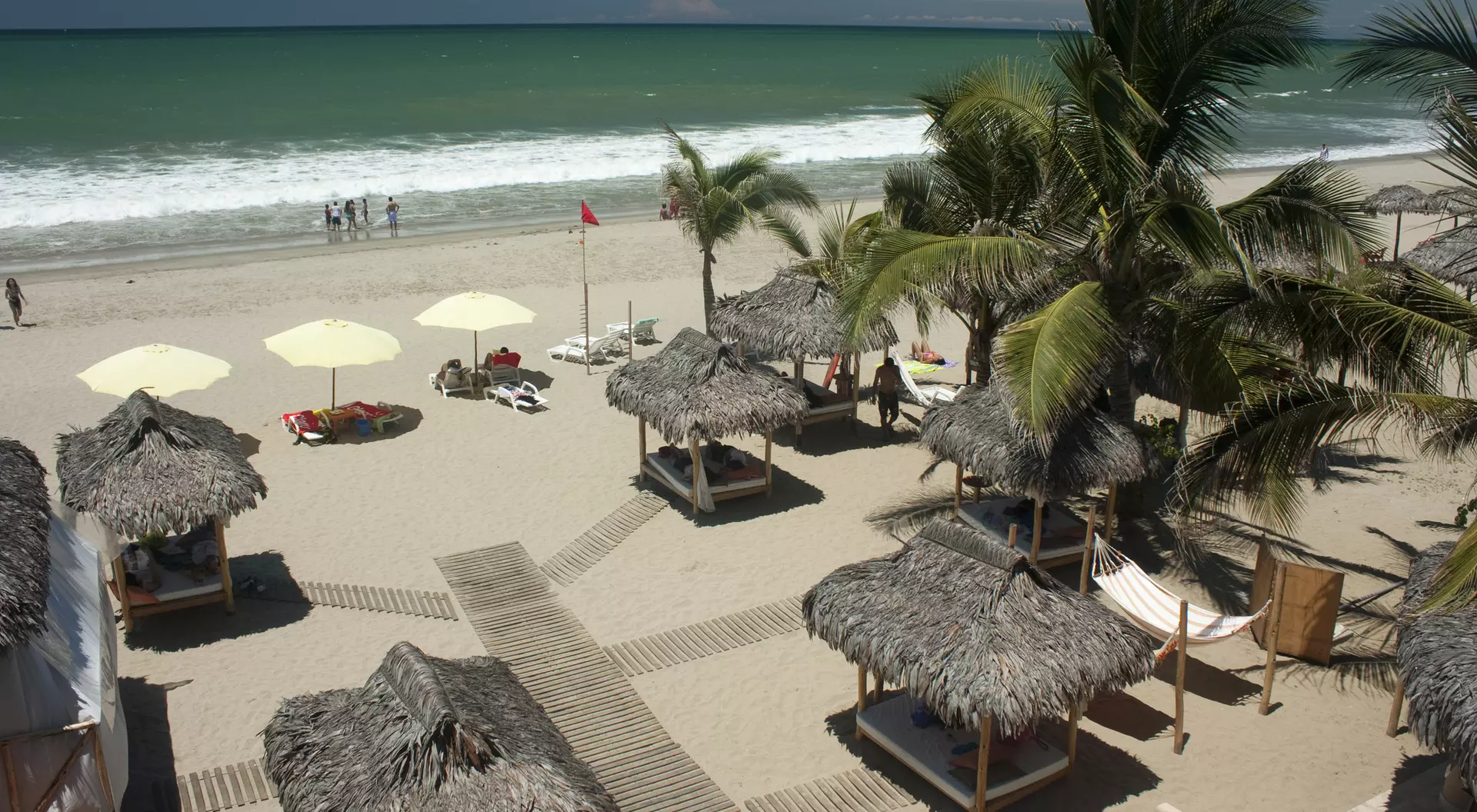 A beach scene with straw huts, palm trees, and umbrellas, featuring gentle waves and people in the background.