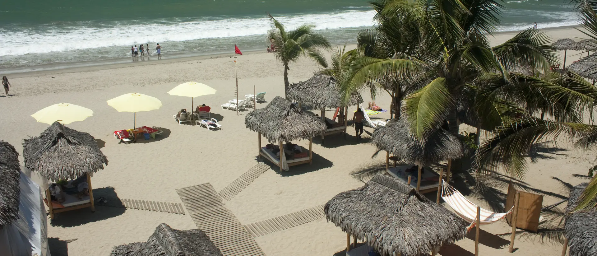 A beach scene with straw huts, palm trees, and umbrellas, featuring gentle waves and people in the background.
