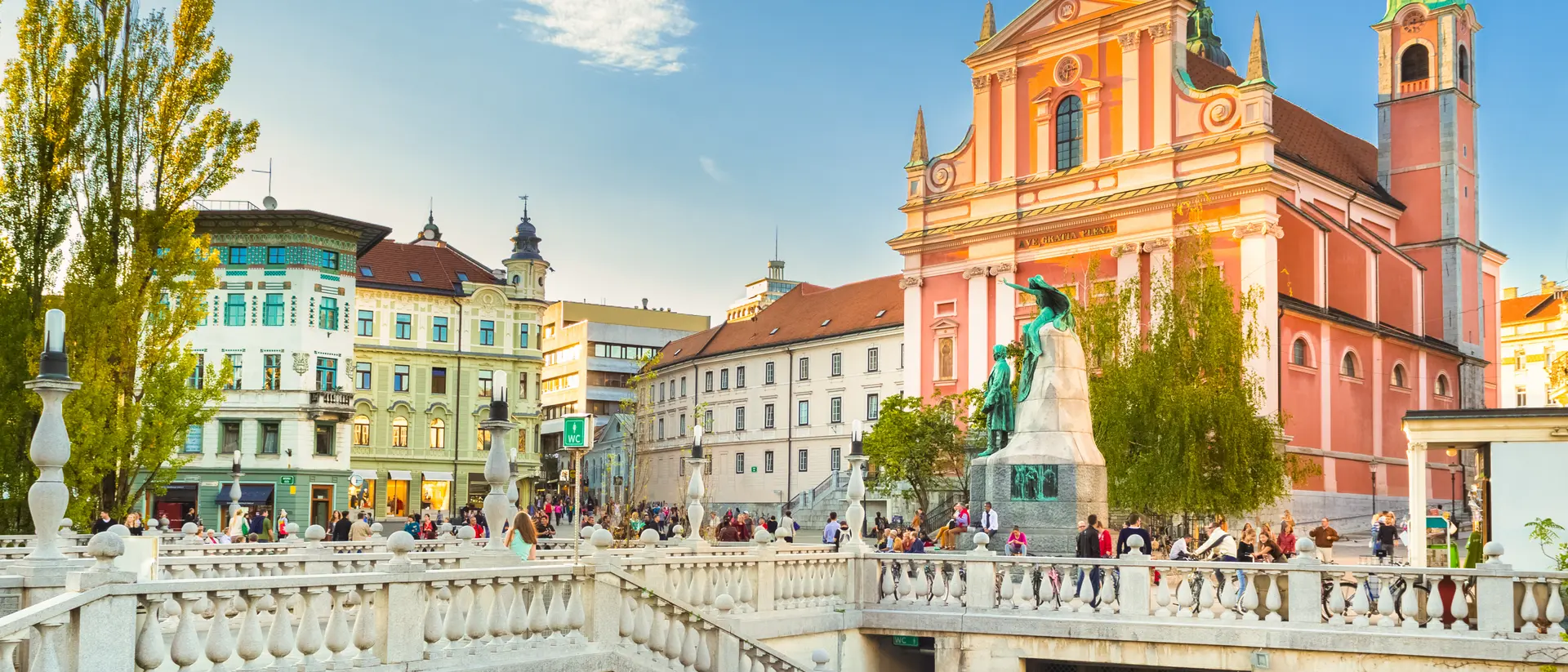 A scenic view of a bustling square with a bridge, colorful buildings, and a church under a clear blue sky.