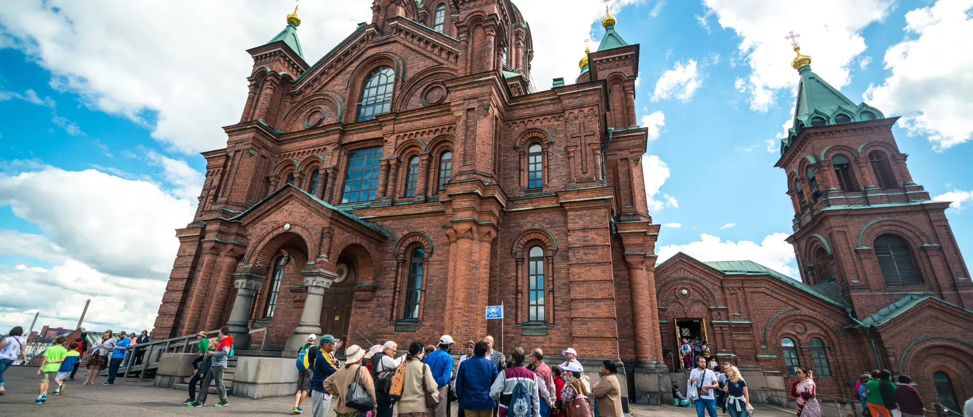 A group of tourists gathers in front of a large, ornate brick church with golden domes against a cloudy sky.