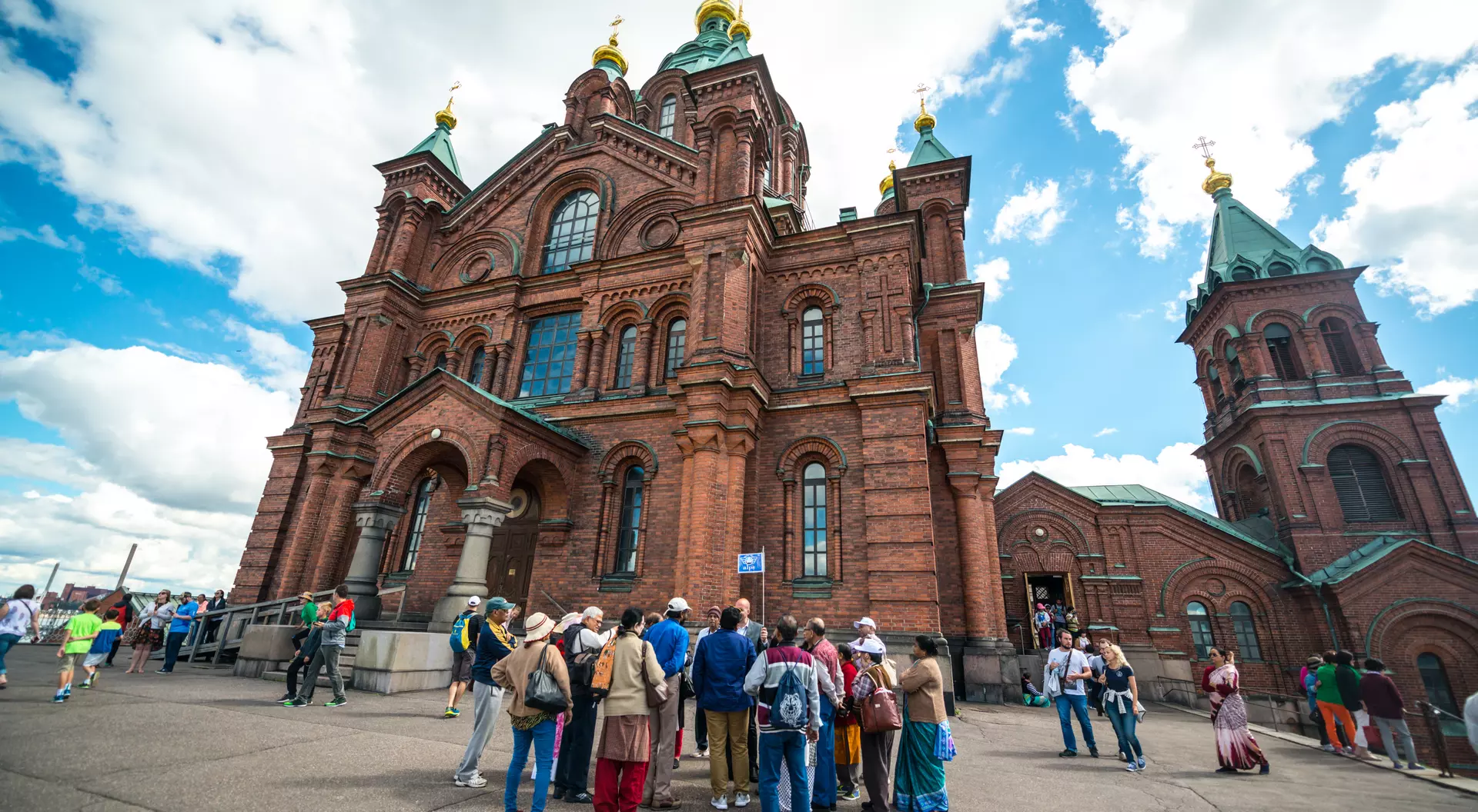 A group of tourists gathers in front of a large, ornate brick church with golden domes against a cloudy sky.