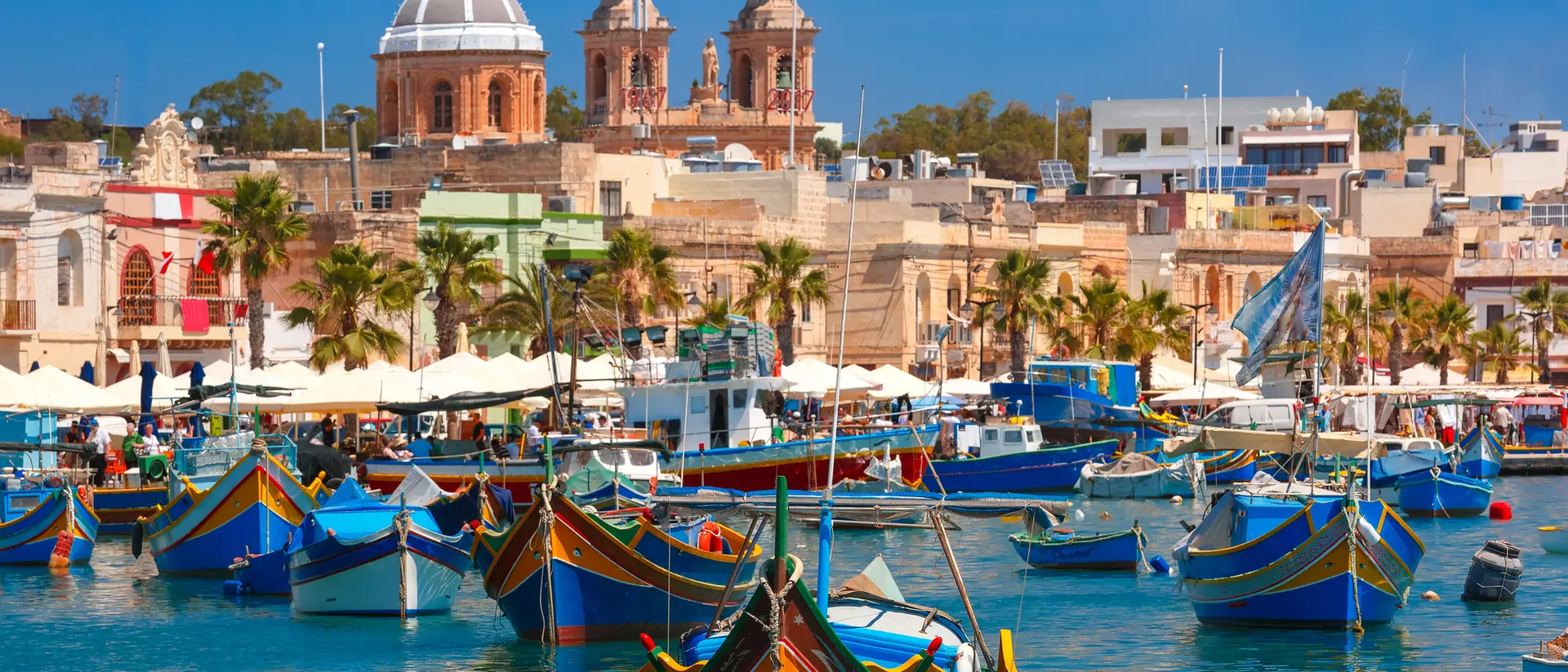 Colorful boats in a vibrant harbor, with historic buildings and palm trees under a bright blue sky.