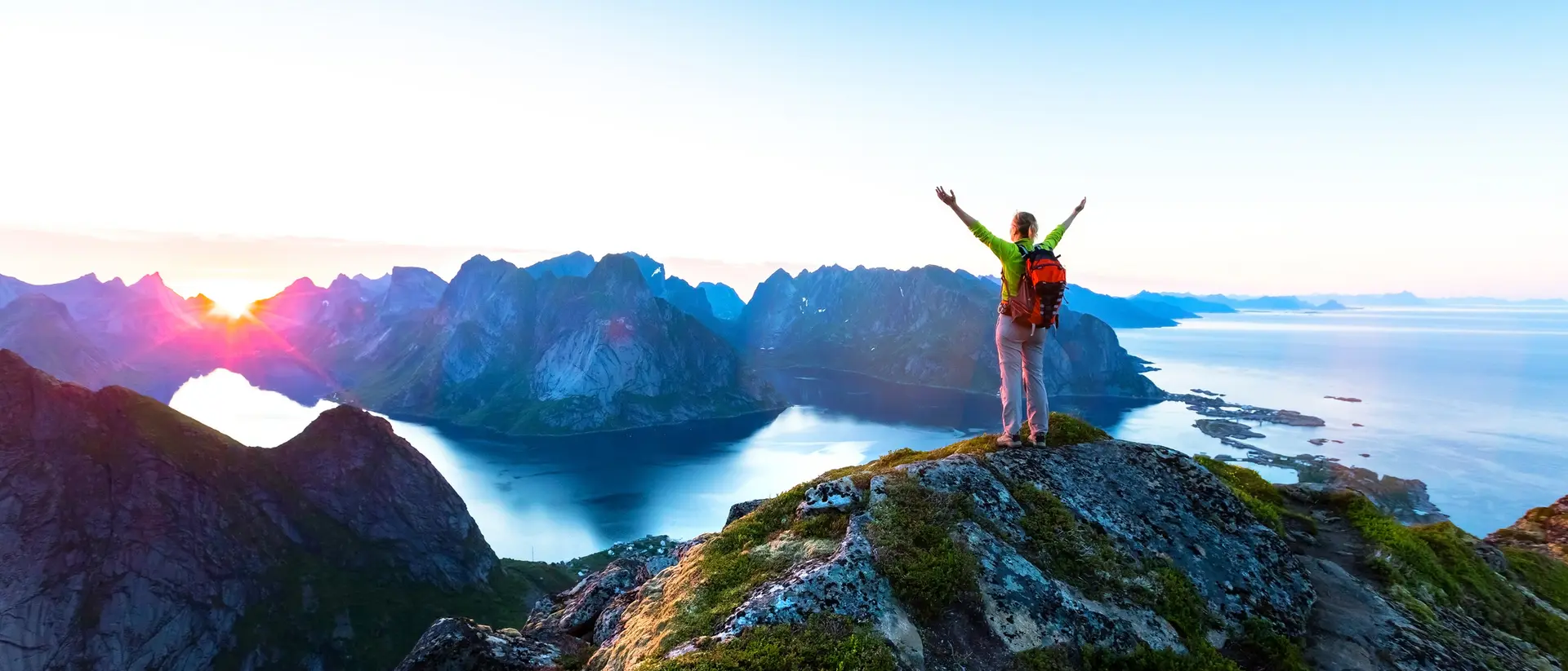 A person with arms raised stands on a rocky peak, overlooking a colorful landscape of mountains and water at sunrise.