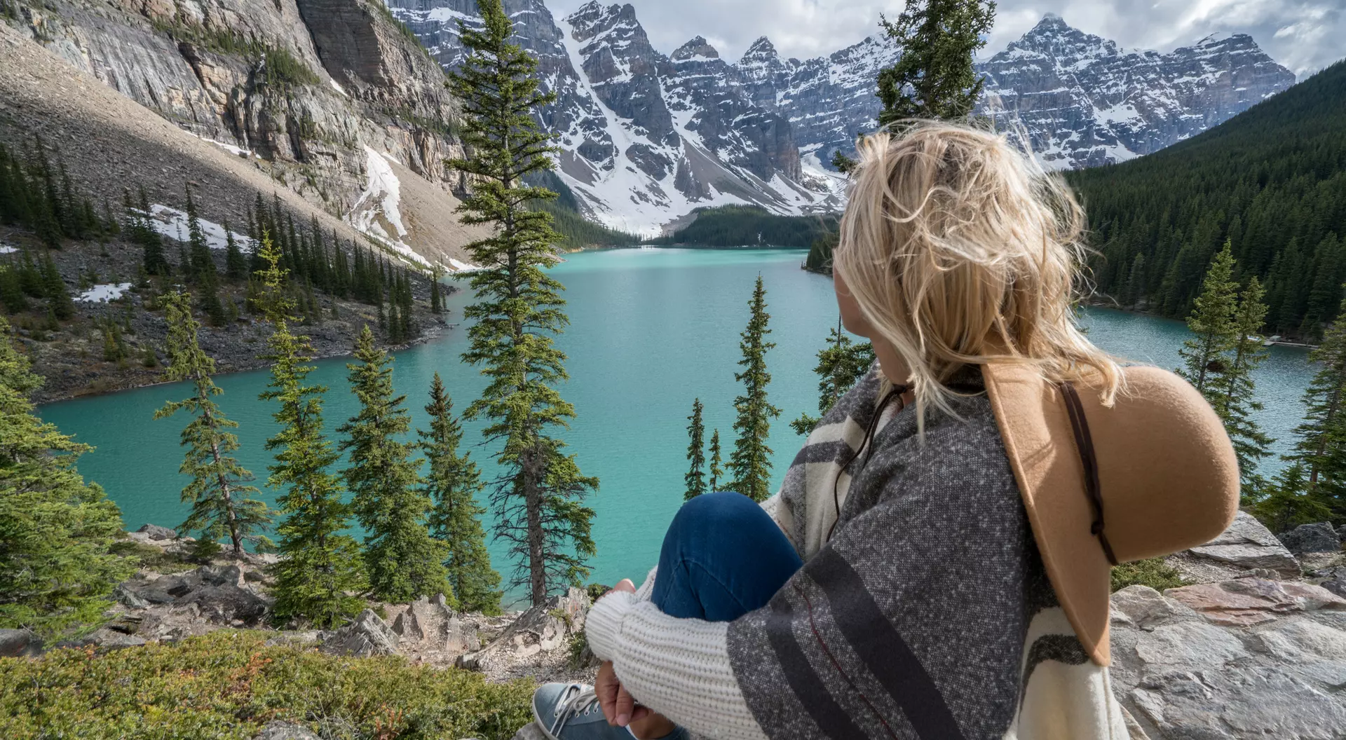 A person sitting on a rock, gazing at a turquoise lake surrounded by mountains and evergreen trees.