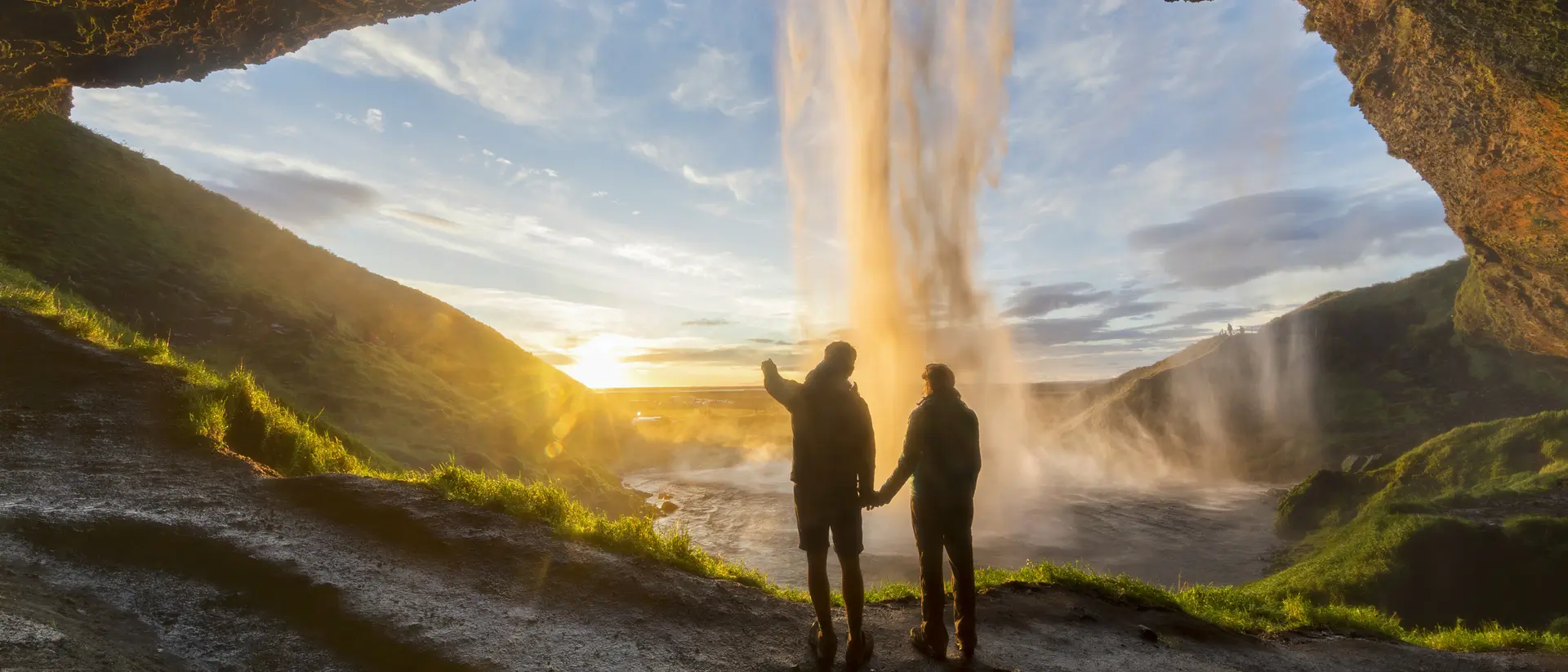 Silhouetted figures stand hand in hand under a waterfall, framed by a rocky cavern and a colorful sunset.