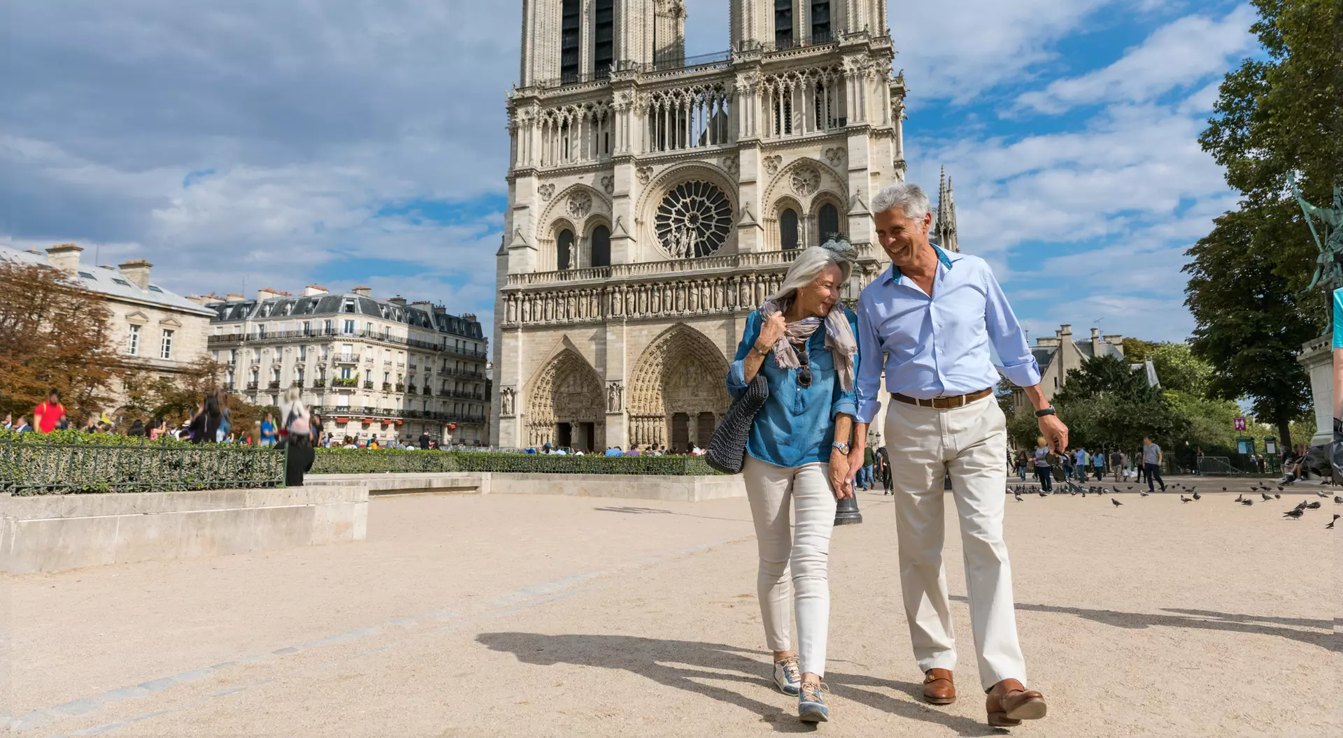 A couple walks hand-in-hand in front of Notre-Dame Cathedral on a sunny day, with people and trees in the background.