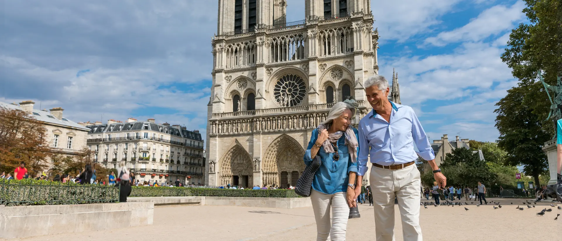 A couple walks hand-in-hand in front of Notre-Dame Cathedral on a sunny day, with people and trees in the background.