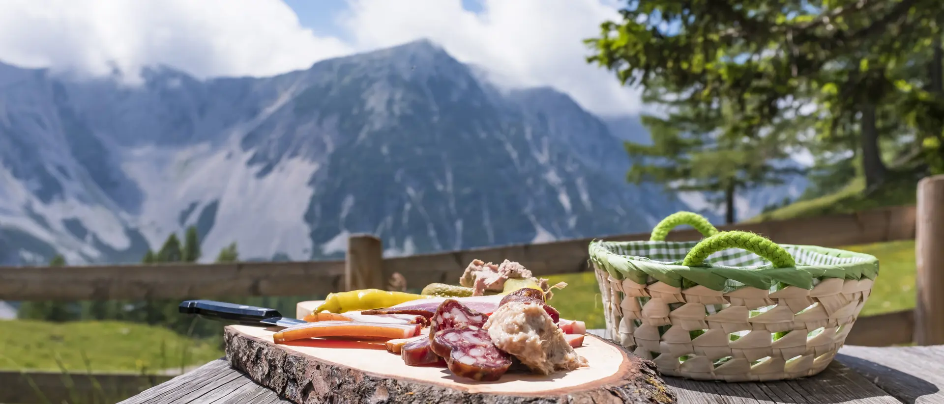 A rustic wooden table holds a charcuterie platter with cured meats and vegetables, set against a scenic mountain backdrop.