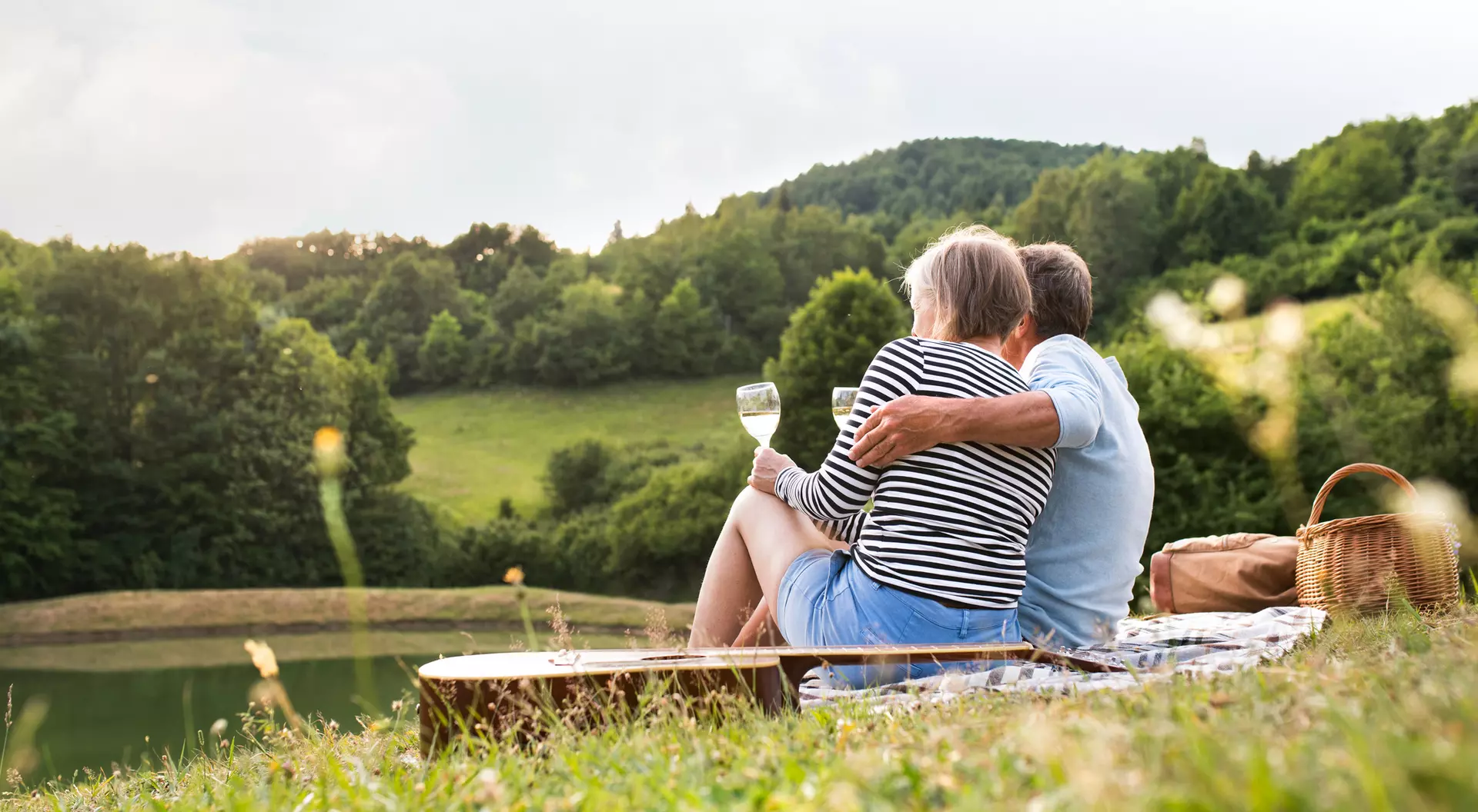 A couple sits together by a serene lake, enjoying a drink and the view of green hills and trees in the background.