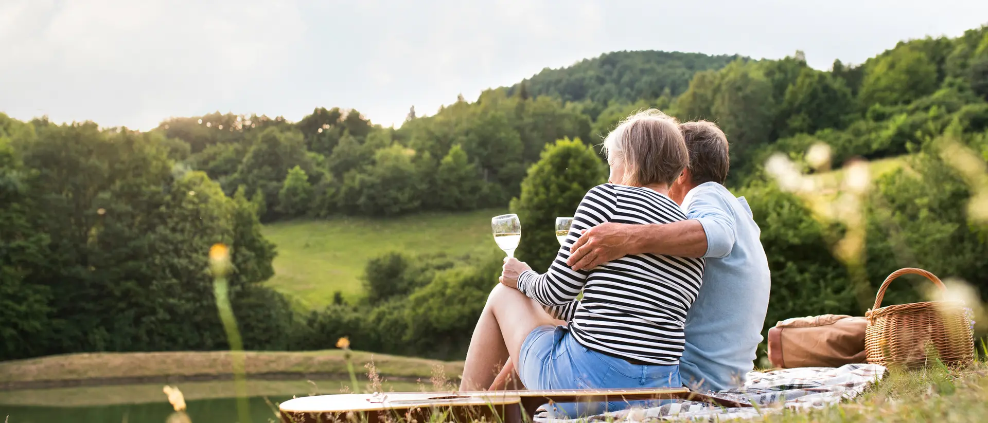 A couple sits together by a serene lake, enjoying a drink and the view of green hills and trees in the background.