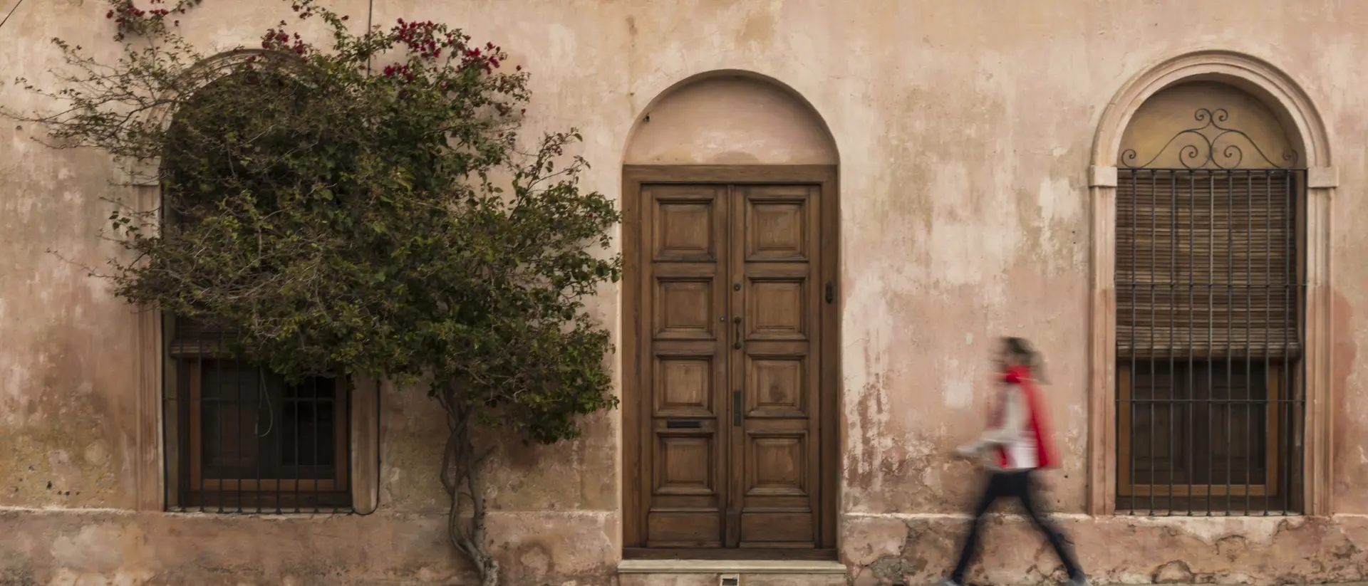 A person walks past a weathered pink building with an arched door and windows, accompanied by a small tree.