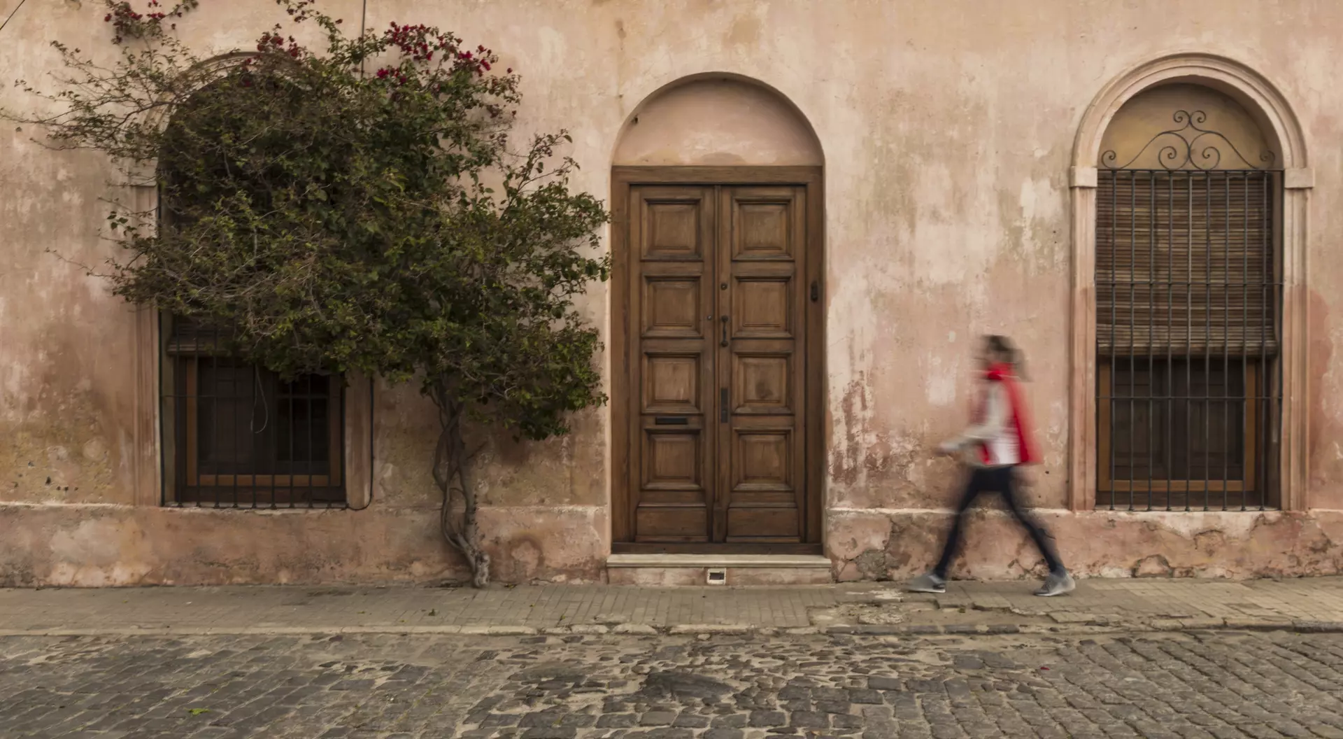 A person walks past a weathered pink building with an arched door and windows, accompanied by a small tree.