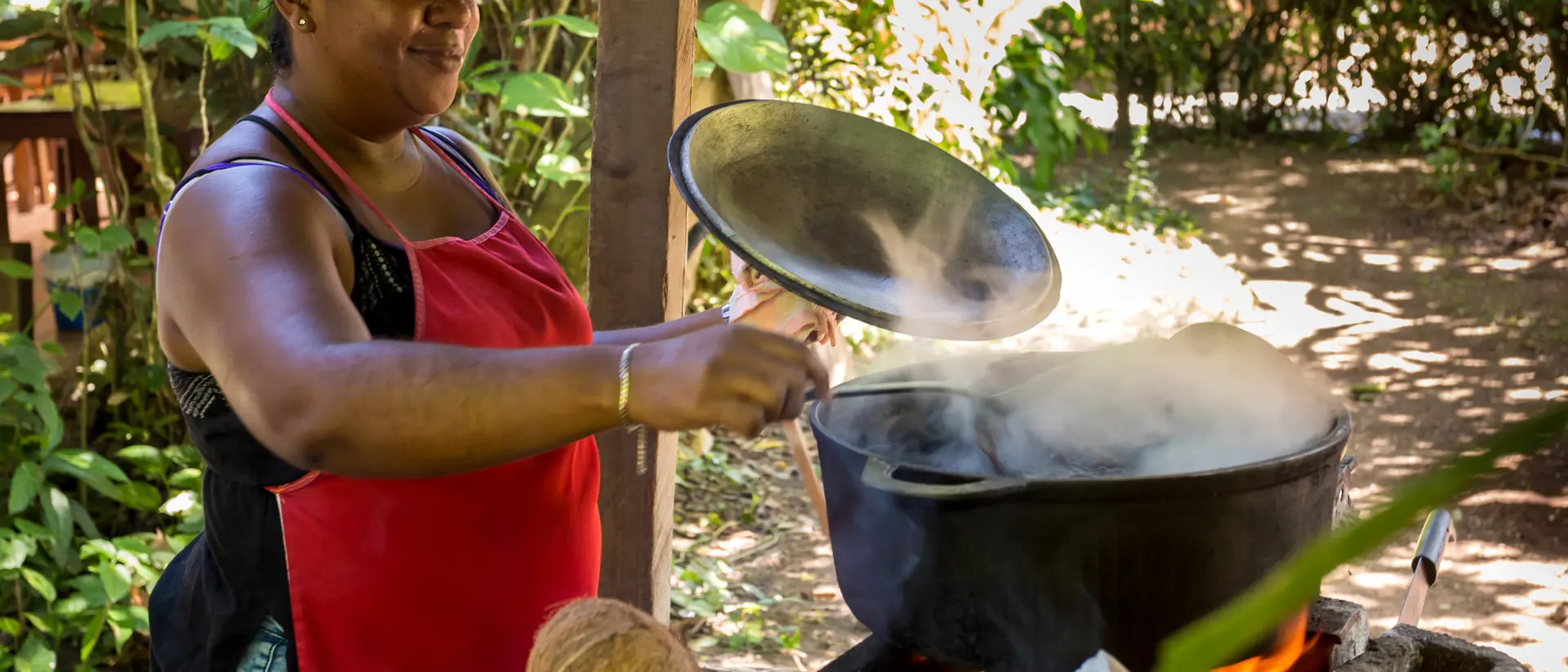 A woman in a red apron stirs a pot over a fire outdoors, surrounded by greenery and coconuts.