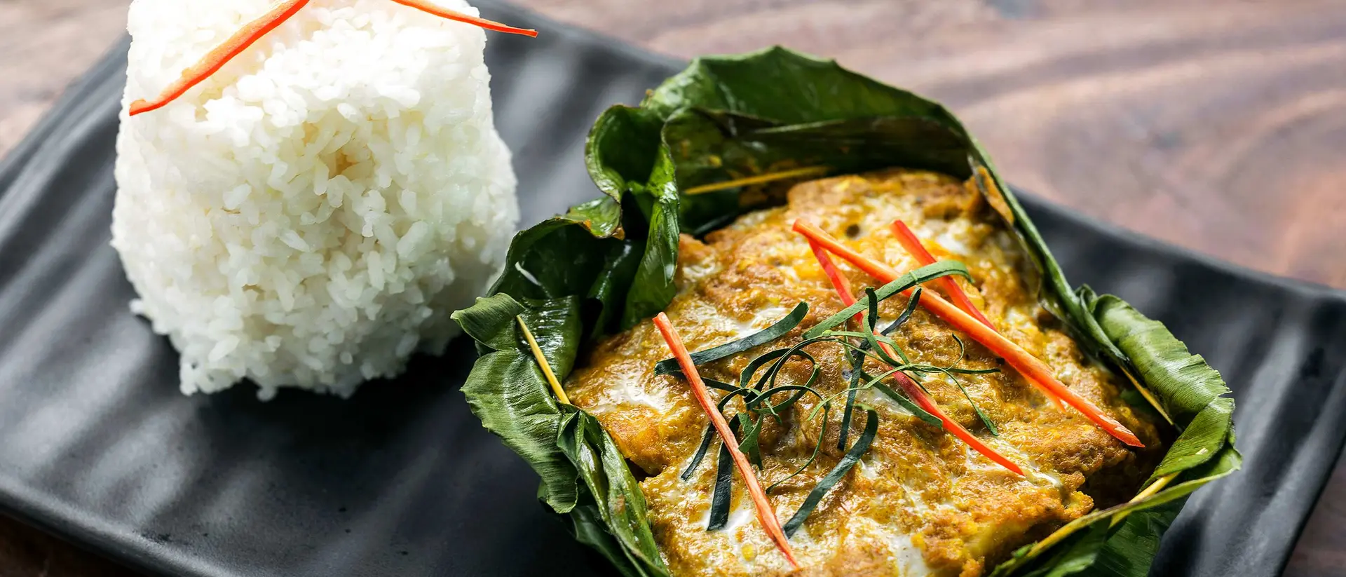A dish featuring fish curry in banana leaves, garnished with herbs and red chilies, served with a side of white rice.
