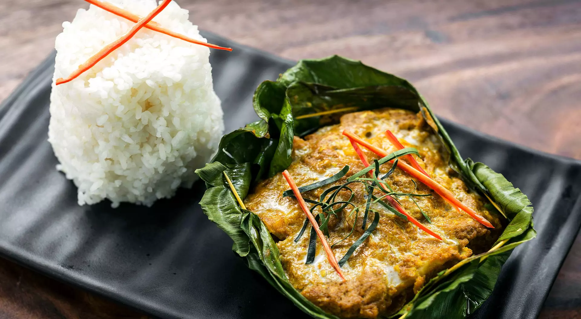 A dish featuring fish curry in banana leaves, garnished with herbs and red chilies, served with a side of white rice.