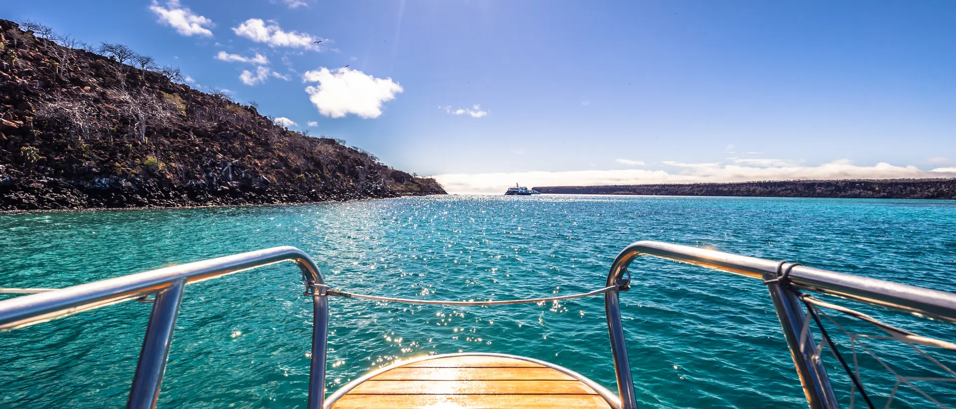 View from the bow of a boat, looking out over turquoise water towards a rocky shoreline under a clear blue sky.