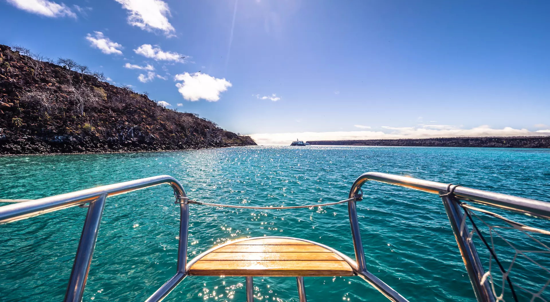 View from the bow of a boat, looking out over turquoise water towards a rocky shoreline under a clear blue sky.