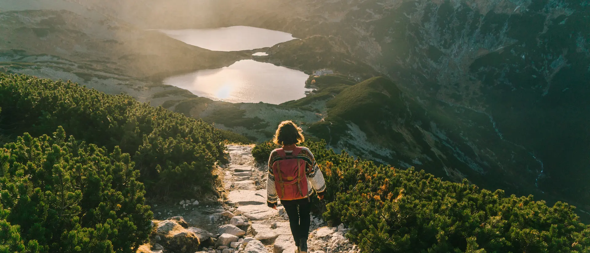 A person walking on a rocky path overlooking two glistening lakes and mountains during sunset.