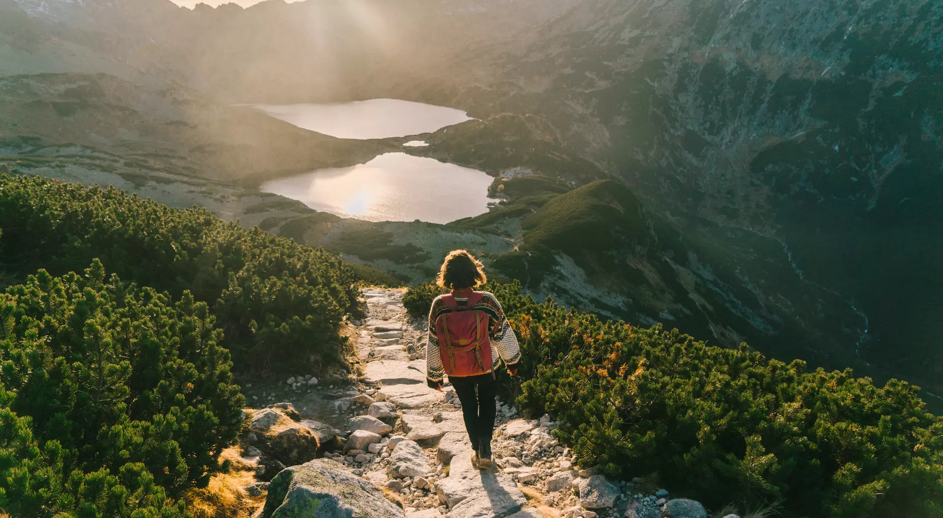 A person walking on a rocky path overlooking two glistening lakes and mountains during sunset.