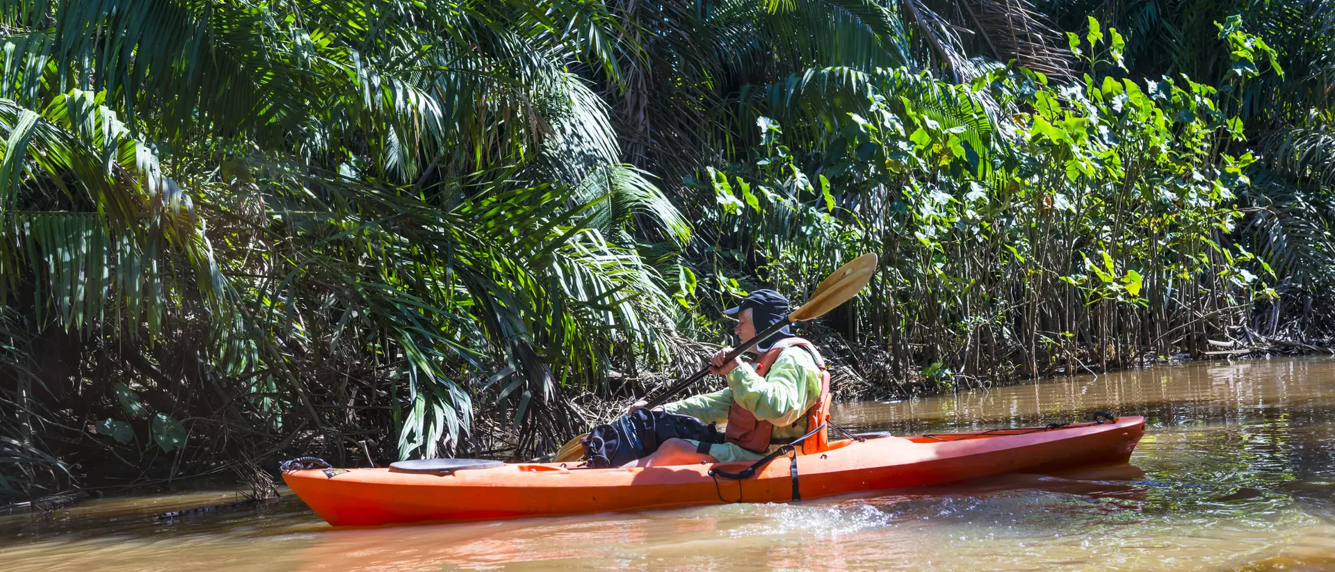 A person paddles a kayak through a lush, green river surrounded by tropical foliage.