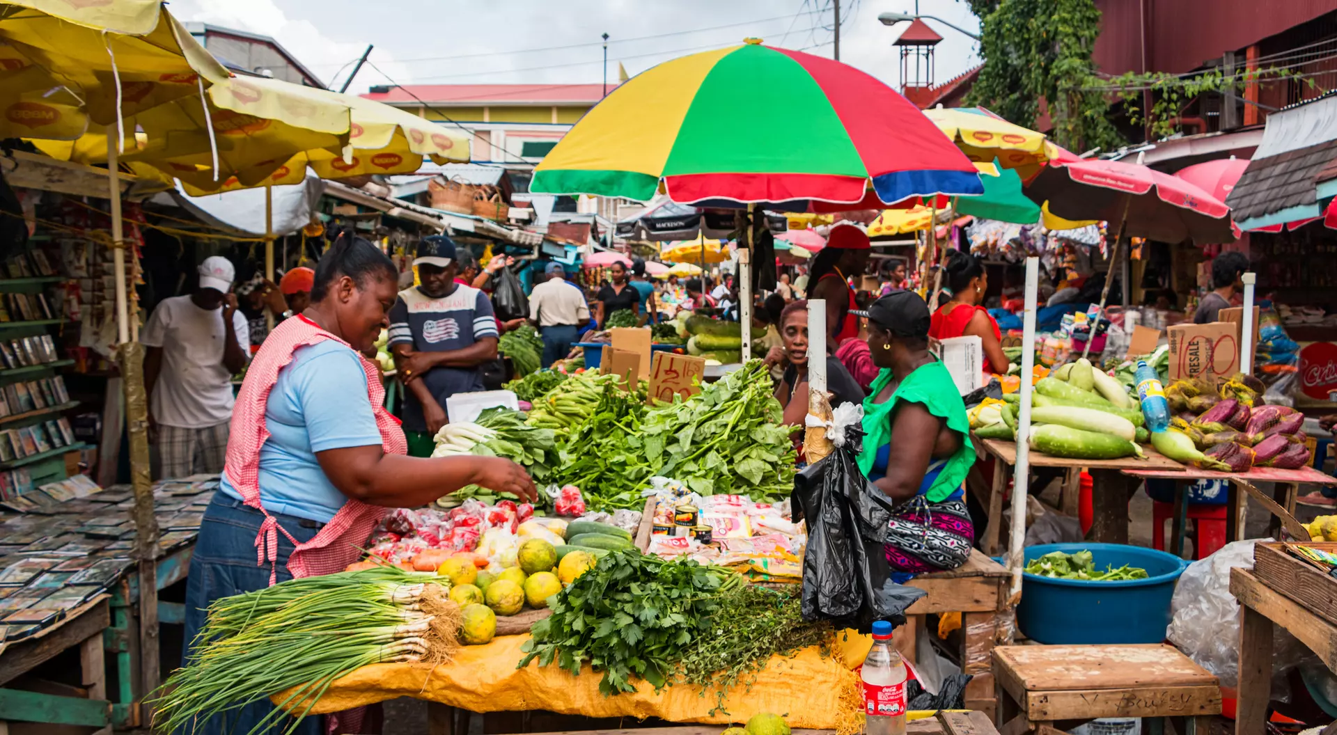 A vibrant market scene featuring vendors selling fresh produce under colorful umbrellas, with shoppers browsing the stalls.