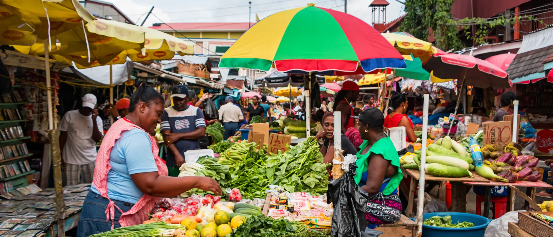 A vibrant market scene featuring vendors selling fresh produce under colorful umbrellas, with shoppers browsing the stalls.