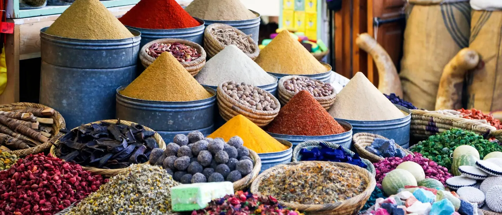 Colorful spices and herbs are displayed in baskets and metal containers at a vibrant market.