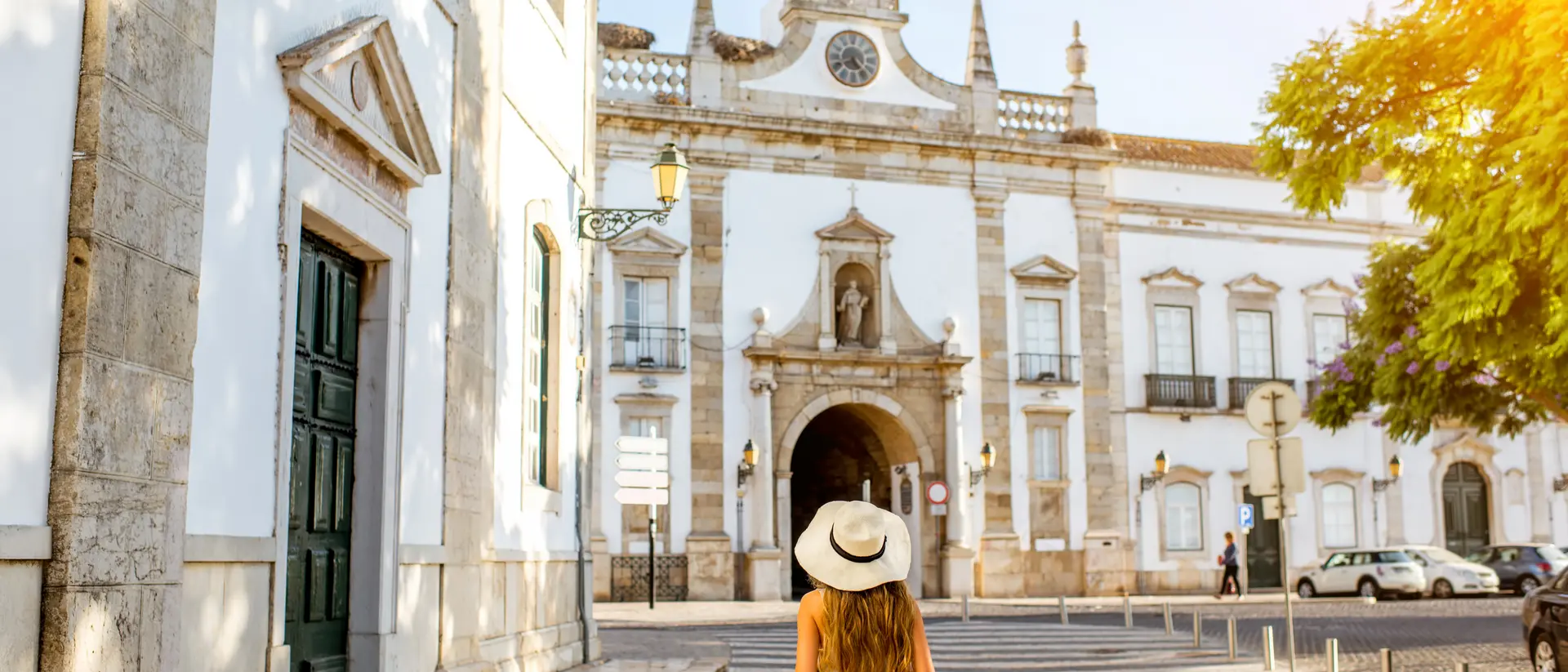 A woman in a hat walks towards a historic building, surrounded by trees and sunlight in a charming street setting.