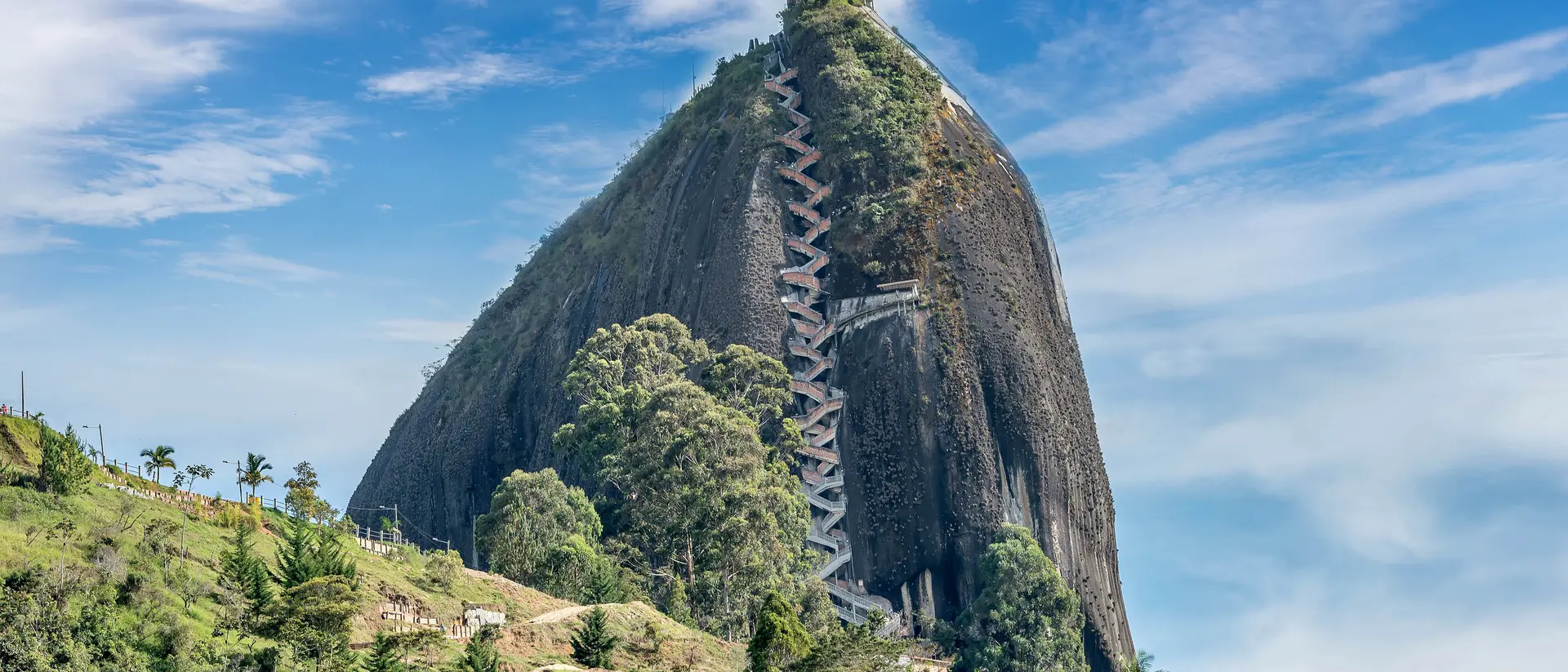 A tall, rocky mountain with a zigzag path and a viewing tower, surrounded by lush greenery and a small houses below.