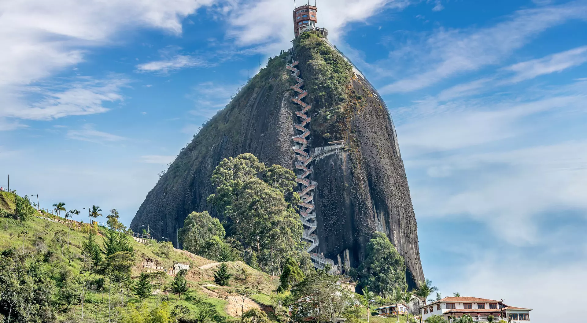 A tall, rocky mountain with a zigzag path and a viewing tower, surrounded by lush greenery and a small houses below.