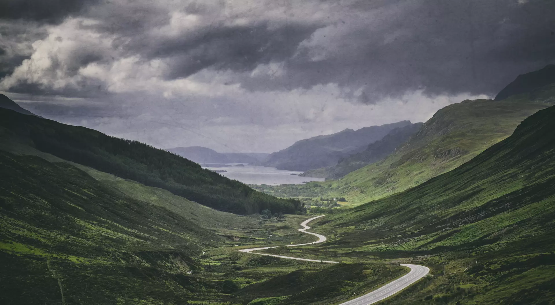 A winding road cuts through lush green valleys under a moody sky, leading to a serene lake in the distance.