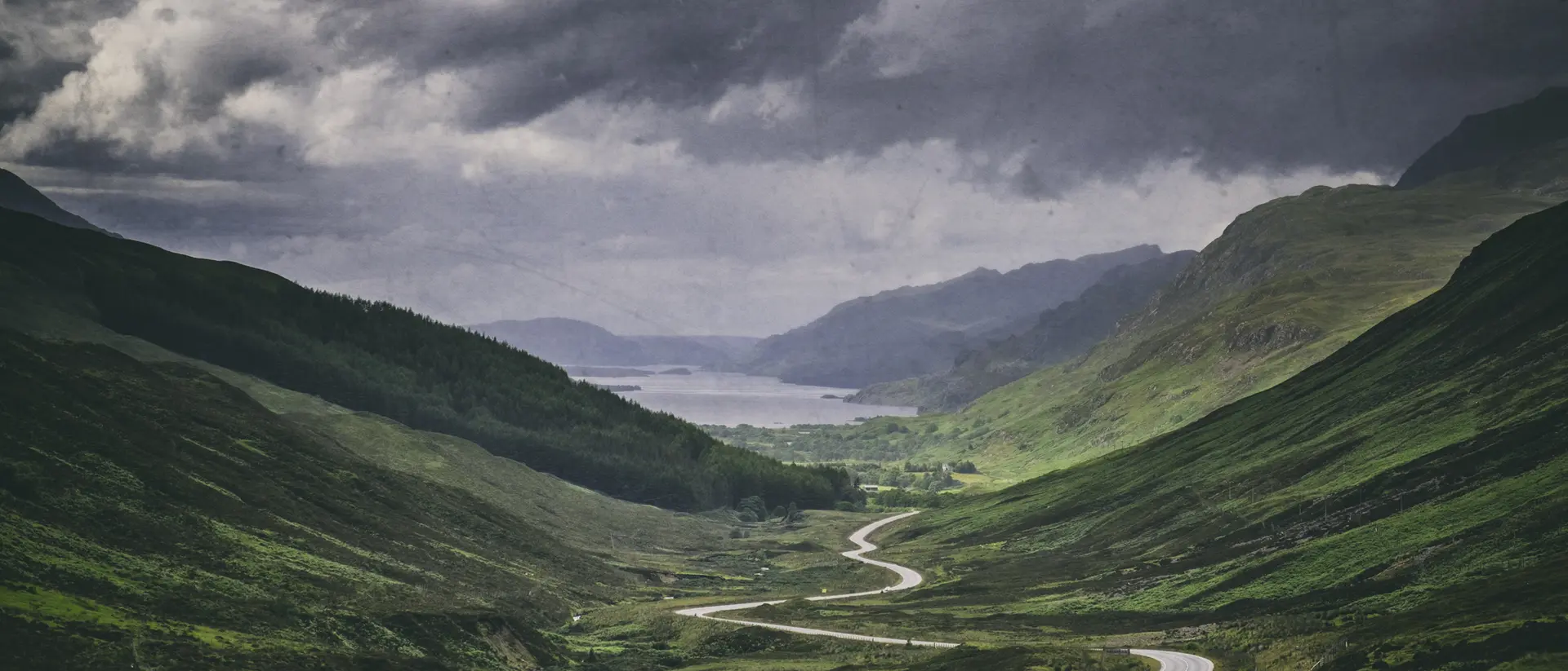 A winding road cuts through lush green valleys under a moody sky, leading to a serene lake in the distance.