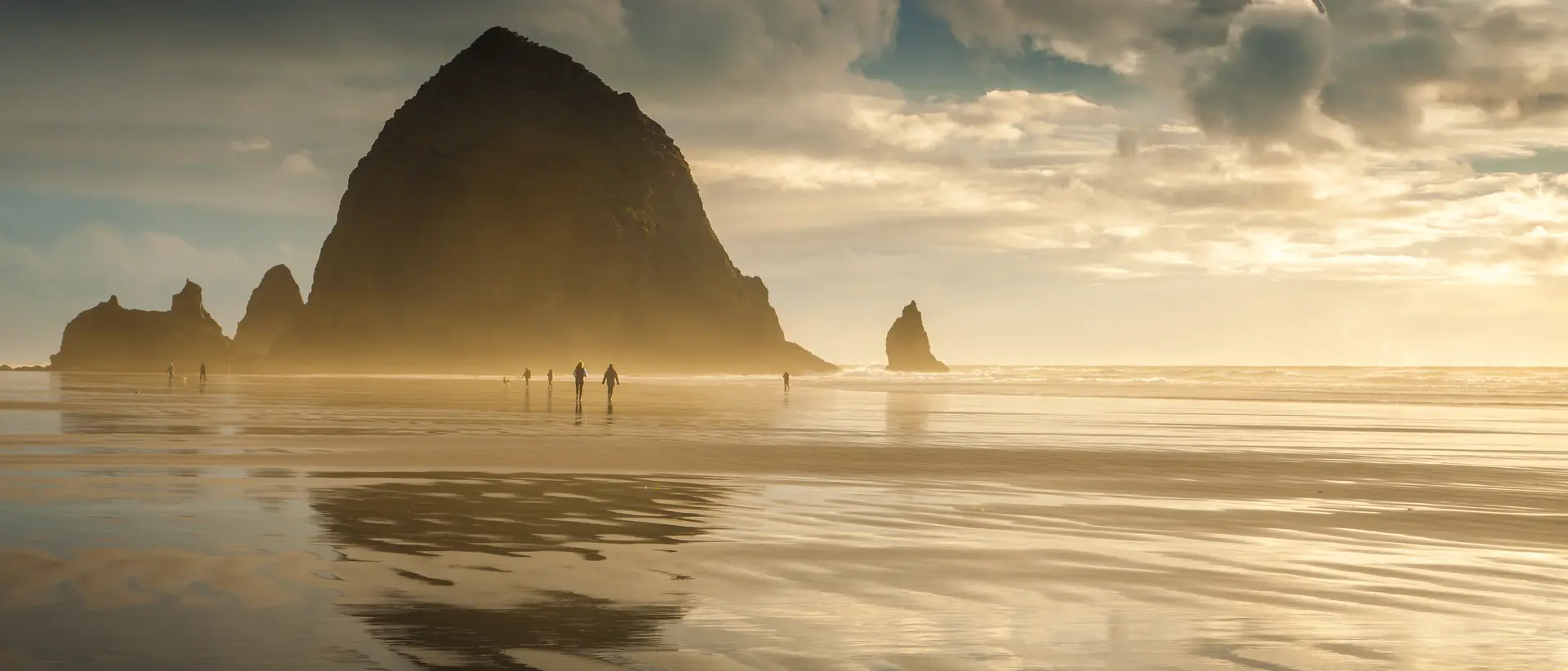 Oregon's Cannon Beach at sunset and in between storms. People can be seen strolling on the beach.