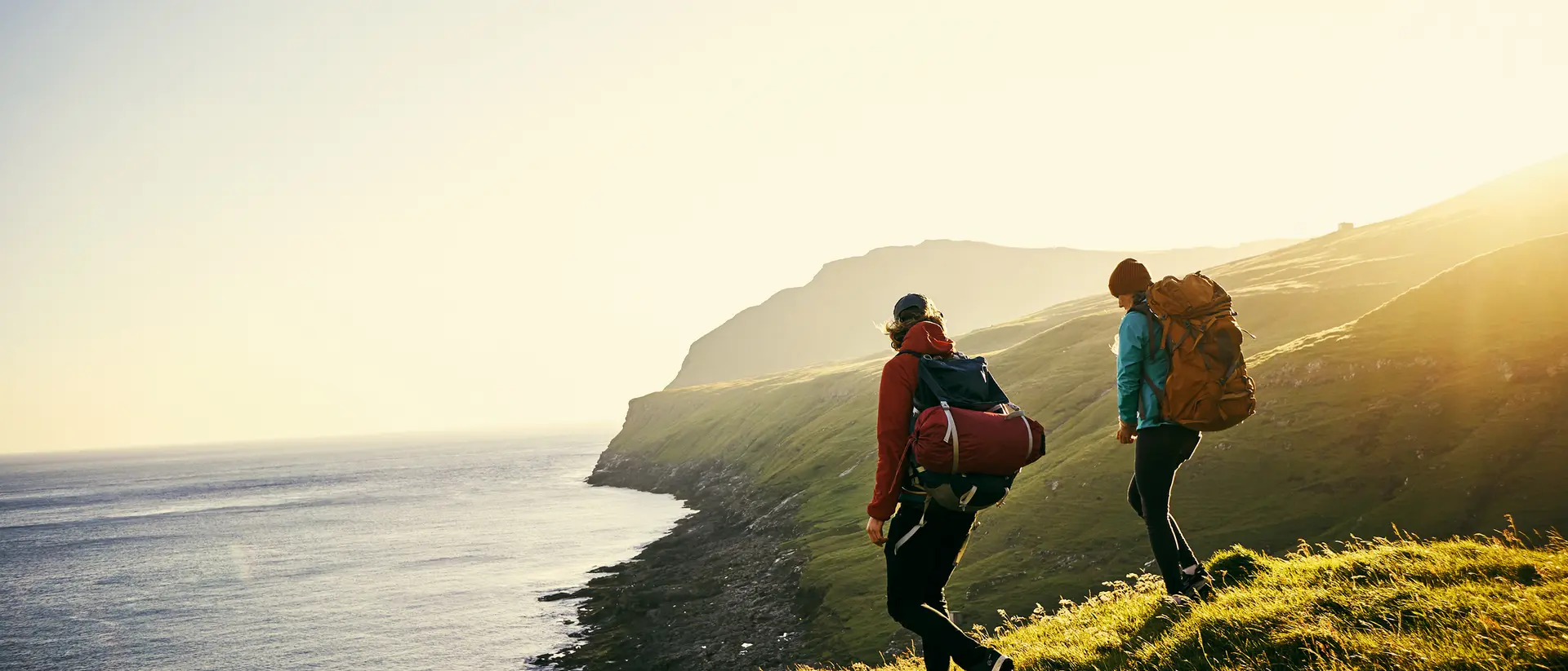 Two hikers with backpacks walk along a grassy hillside, overlooking a calm sea under a bright sunset.