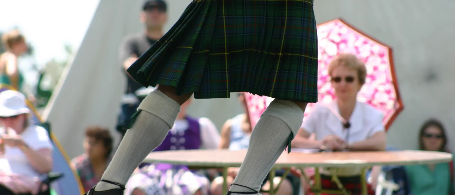 A dancer in a kilt performs on stage, while an audience watches, some under a colorful umbrella.